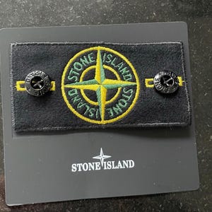 Puede incluir: Un parche de tela rectangular negro con un logotipo de brújula bordado en amarillo y verde y el texto STONE ISLAND. Dos botones negros están unidos al parche. El parche está montado sobre una tarjeta negra con el texto blanco STONE ISLAND.