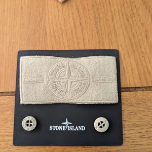 May include: A rectangular beige fabric patch featuring an embroidered Stone Island compass logo and text. Below the patch, a black display card holds two beige buttons, each with the Stone Island name and logo embossed on the surface.