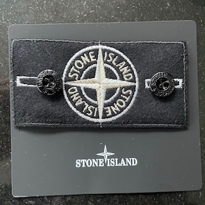Puede incluir: Un parche rectangular de tela negra con el logotipo de la brújula Stone Island bordado en blanco y gris. El parche está sujeto a una tarjeta de soporte gris oscuro con dos botones negros, cada uno grabado con el nombre de la marca Stone Island.