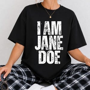 May include: A black short-sleeved t-shirt featuring the white distressed-style text I AM JANE DOE printed on the front. The shirt is paired with black and white plaid pants and white sneakers, with a pair of sunglasses held in one hand.