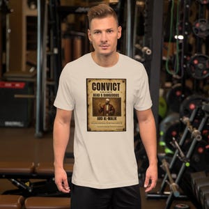 May include: A cream-colored short-sleeve t-shirt featuring a rectangular graphic print that resembles a vintage wanted poster. The text reads CONVICT, DEAD & DANGEROUS, and ABD AL-MALIK, centered above an illustration of a person in traditional attire.