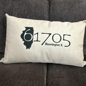 Könnte beinhalten: Ein beiges Kissen mit einer schwarzen Umrandung des Bundesstaates Illinois und dem aufgedruckten Text "61705 Bloomington, IL".