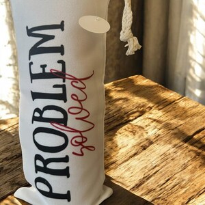 Puede incluir: Bolsa de vino de lona blanca con las palabras "PROBLEM solved" impresas en negro y rojo. La bolsa tiene un cierre de cordón y una pequeña etiqueta adjunta. La bolsa está sobre una superficie de madera.