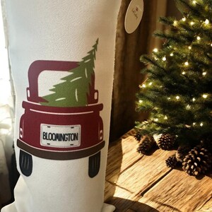 Puede incluir: Un calcetín navideño blanco con un diseño de camión rojo que transporta un árbol de Navidad verde. La matrícula del camión dice "BLOOMINGTON". El calcetín está sobre una superficie de madera cerca de un árbol de Navidad iluminado.