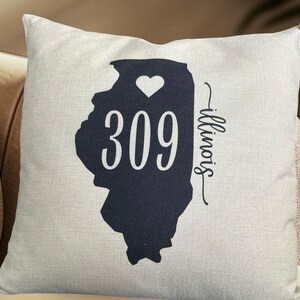 May include: A square cushion featuring a navy blue outline of the state of Illinois, a white heart, and the number 309 in white. The word "Illinois" is written in cursive. The cushion is on a beige background.