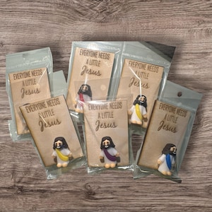 May include: Several small Jesus figurines are packaged in clear plastic. Each figurine is paired with a wooden card that reads "EVERYONE NEEDS A LITTLE Jesus." The figurines have different colored robes.