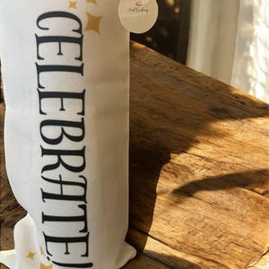 Puede incluir: Un artículo de tela blanca con la palabra "CELEBRATE" impresa verticalmente en negro, acompañada de gráficos de estrellas doradas. Se adjunta una pequeña etiqueta redonda con texto. El artículo está sobre una superficie de madera.