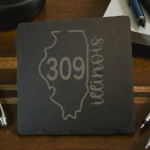 May include: A square, dark gray slate coaster with the outline of the state of Illinois etched into the surface. The number "309" is inside the state outline, and the word "Illinois" is written vertically along the right side.
