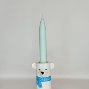 Könnte beinhalten: Ein weißer Kerzenhalter aus Keramik in Form eines Eisbärenkopfes mit einem blau gepunkteten Schal, der eine hohe, spitz zulaufende hellminzgrüne Kerze hält. Der Bär hat kleine Ohren, schwarze Augen, eine schwarze Nase und aufgemalte rosa Wangen.