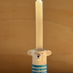 Può includere: Un portacandele in ceramica a forma di orso con viso bianco, piccole orecchie e corpo a righe blu e bianche. L'orso presenta naso e occhi neri dipinti e un piccolo cuore rosso sulla guancia. È inserita una candela affusolata color bianco sporco.