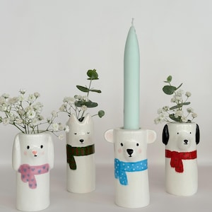 May include: Four white ceramic animal-shaped vases, each featuring a painted scarf. Three vases hold sprigs of baby's breath and eucalyptus, while the central bear-shaped vase holds a tall, light blue taper candle. Each piece has a unique face.