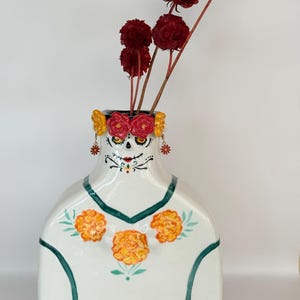 Op de afbeelding: Een witte keramische vaas in de vorm van een torso, voorzien van een geschilderd suikerschedelgezicht met gele ogen en zwarte details. De vaas is versierd met een krans van rode en gele bloemen, bungelende bloemenoorbellen en oranje goudsbloemmotieven.