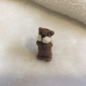 May include: A small brown and white felt animal figurine of an otter. The otter is standing upright with its paws together and its head tilted slightly to the side. The otter has a brown body, white face, and black eyes.