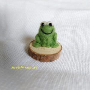 May include: A small green felt frog with white eyes sits on a wooden log. The frog is smiling and has a black mouth.