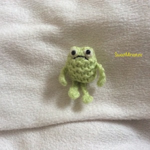 May include: A small green knitted frog with black eyes and a frown. The frog has four limbs and is sitting on a white surface.