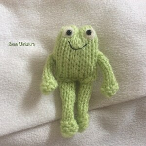 May include: A green knitted frog toy with white eyes and a smile. The frog is standing on a white surface.