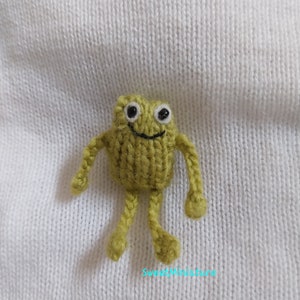 May include: A small knitted green frog with black eyes and a smile. The frog has long arms and legs.