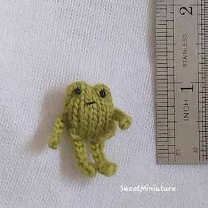 May include: A small, green knitted frog with black eyes. The frog is about 1 inch tall and is sitting on a white surface next to a ruler.