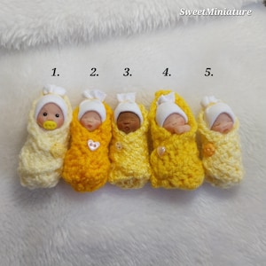 May include: Five miniature baby figurines wrapped in yellow crocheted blankets and wearing white knit hats, numbered one through five. Each figurine has a distinct facial expression and skin tone, with small decorative buttons on the blankets.
