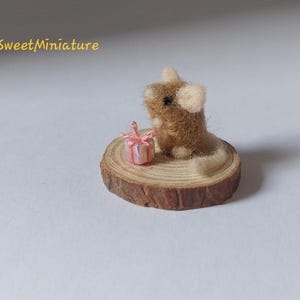 May include: A miniature felted mouse figurine with brown fur and a white tail, sitting on a small wooden slice. Beside the mouse is a tiny pink gift box tied with a pink ribbon. The text "SweetMiniature" is in the upper left corner.