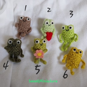 May include: Six crocheted frog plushies in various colors, including green, brown, and yellow. The frogs are numbered 1 through 6. The plushies are handmade and have a sweet, whimsical style.