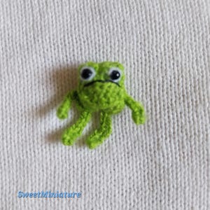 May include: A small, handmade green crochet frog with black eyes and a black mouth. The frog has small arms and legs and is positioned on a white knitted background. The text "SweetMiniature" is in the bottom left corner.
