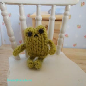 May include: A small, green knitted frog toy with black eyes sitting on a white chair. The frog has a simple, cartoon-like design.