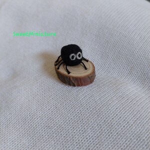 May include: A small black felt spider with white eyes sits on a small wooden log. The spider is about 1 inch in diameter.