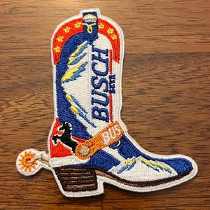 May include: An embroidered patch shaped like a cowboy boot featuring white, blue, and red colors. The text BUSCH BEER is stitched vertically in blue. It includes a black horse silhouette, a yellow spur, and a small orange strap with BUSCH text.