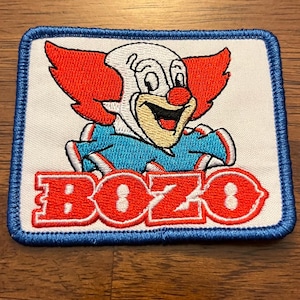 May include: A rectangular embroidered patch featuring a white background with a blue border. The design depicts a clown with bright red hair, a red nose, and a blue collar. The word BOZO is embroidered in bold red letters at the bottom.