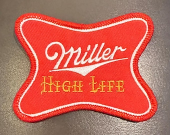 May include: A red embroidered patch with a white border shaped like a stylised cross. The patch features the white cursive text Miller above the yellow serif text HIGH LIFE. The embroidery has a textured finish with a distinct red stitched edge.