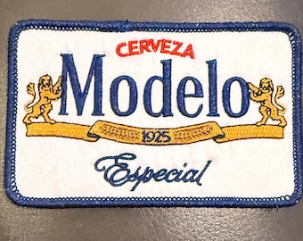 May include: A rectangular embroidered patch with a white background and a blue border. It features the text CERVEZA Modelo in blue, with two gold lions on either side. A gold ribbon banner displays the year 1925, and the word Especial is below.