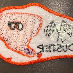 May include: An embroidered patch with a white background and a bright orange stitched border. It features a stylised orange tornado graphic with black sunglasses, two crossed chequered racing flags, and the word Duster in black text.
