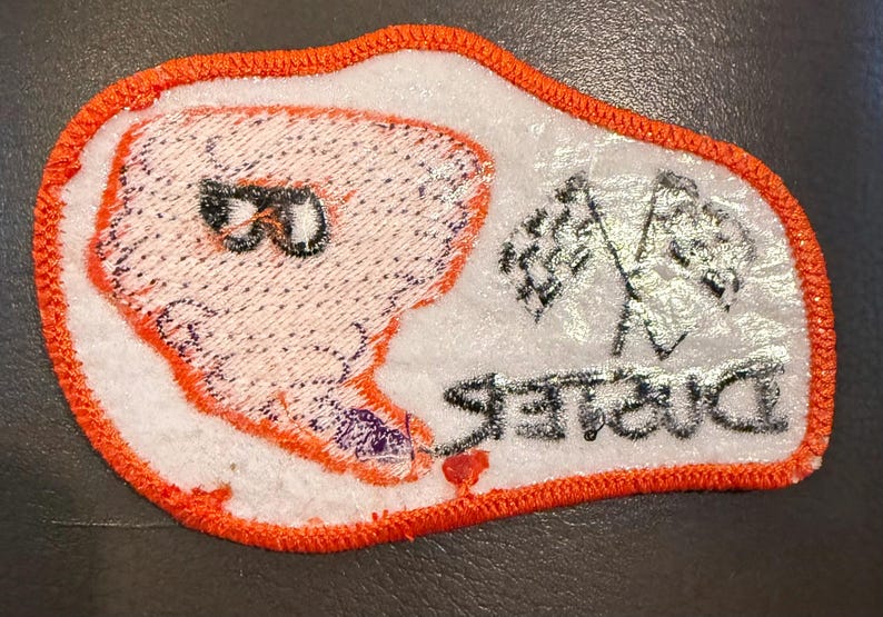 May include: An embroidered patch with a white background and a bright orange stitched border. It features a stylised orange tornado graphic with black sunglasses, two crossed chequered racing flags, and the word Duster in black text.