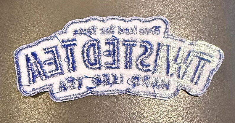 May include: A white embroidered patch with a scalloped edge featuring the blue text TWISTED TEA in a bold, stylised font. The design includes smaller blue text above and below the main brand name, all rendered in a clean, stitched thread finish.