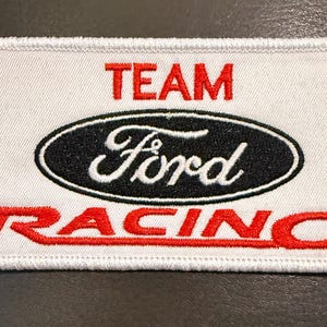 May include: A rectangular embroidered patch with a white background and a stitched border. The text TEAM is in red above a black oval containing the white Ford logo. The word RACING is embroidered in red letters across the bottom section.