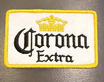 May include: A rectangular embroidered patch featuring a white background with a yellow border. The text Corona Extra is stitched in black, centred below a yellow embroidered crown. The design is clean and clearly defined with visible stitching.