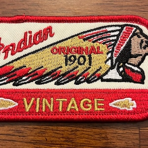 May include: A rectangular embroidered patch with a red border. The design features the red text Indian, a yellow and red feathered headdress, and the black text ORIGINAL 1901. Below, a red section contains the gold text VINTAGE with two small gold arrowheads.