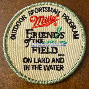 May include: A circular embroidered patch with a tan background and a green stitched border. It features the text "OUTDOOR SPORTSMAN PROGRAM", a red "Miller" logo, and the text "FRIENDS OF THE FIELD" with a small green and blue graphic.