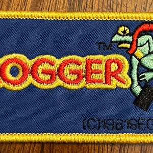 May include: A rectangular embroidered patch with a blue background and a yellow stitched border. It features the red text FROGGER with a yellow outline, a light green frog character, and the black text (C)1981SEGA at the bottom right.