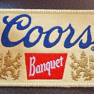 May include: A rectangular embroidered patch with a cream-colored background and a stitched border. The text Coors is embroidered in blue, with the word Banquet in white on a red trapezoid, flanked by two gold-colored lion emblems on each side.