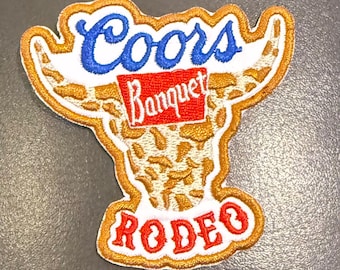 May include: An embroidered patch featuring a stylised bull head design in gold and white. The text Coors is in blue, Banquet is in white on a red background, and RODEO is in red at the bottom. The patch has a gold border and white backing.