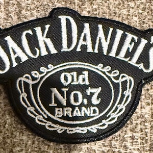 May include: A black embroidered patch featuring the white text JACK DANIEL'S in an arched font at the top. Below, a central oval contains the white text Old No.7 BRAND surrounded by a decorative white stitched border on a black fabric background.