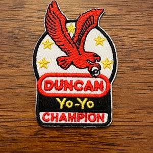 May include: A fabric patch featuring a red eagle graphic above the text DUNCAN YO-YO CHAMPION. The design includes five yellow stars on a white background, with red and black lettering, all bordered by a thick black stitched edge.