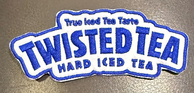 May include: An embroidered patch featuring the blue and white Twisted Tea logo with the text True Iced Tea Taste and Hard Iced Tea. The patch has a scalloped white border and bold blue lettering on a white background with a blue outline.