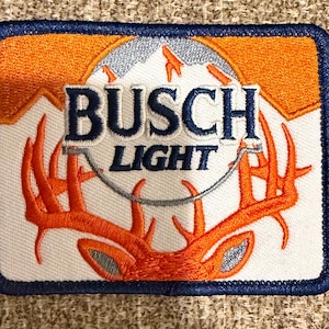 May include: A rectangular embroidered patch featuring the Busch Light logo in dark blue text over a white circular background. The design includes an orange mountain silhouette at the top and a large orange deer antler graphic at the bottom.