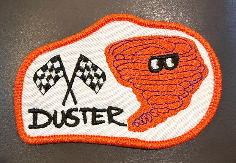 May include: An embroidered patch with a white background and an orange border. It features two crossed black and white chequered flags, the word DUSTER in black text, and an orange cartoon tornado character with black eyes.