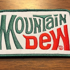 May include: A rectangular embroidered patch with a white background and a dark green stitched border. The text Mountain is embroidered in dark green, and the text Dew is embroidered in bright red, featuring a small registered trademark symbol.