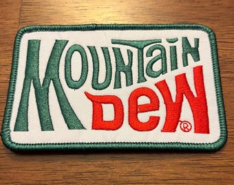 May include: A rectangular embroidered patch with a white background and a dark green stitched border. The text Mountain is embroidered in dark green, and the text Dew is embroidered in bright red, featuring a small registered trademark symbol.