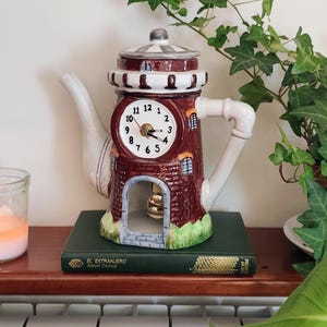 Peut inclure: Une horloge en céramique en forme de théière rappelant un phare en briques avec un bec et une anse blancs. Le cadran affiche des chiffres et des aiguilles noirs. Un petit pendule est suspendu dans la base arquée. Le livre porte le titre El Extranjero.